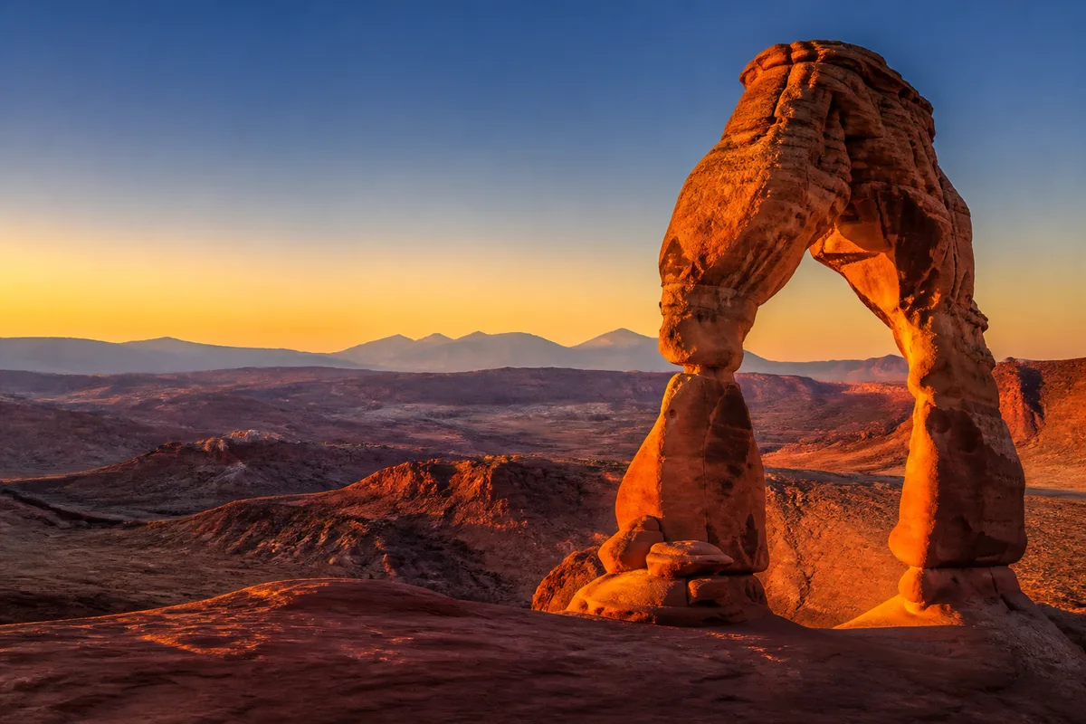 Arche naturelle de grès rouge se détachant sur un ciel clair au coucher du soleil, désert de roche en contrebas