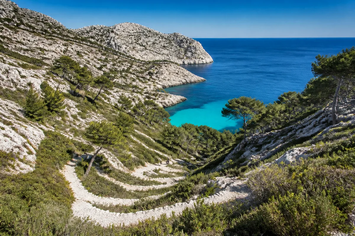 Sentier calcaire étroit descendant vers une crique turquoise bordée de pins